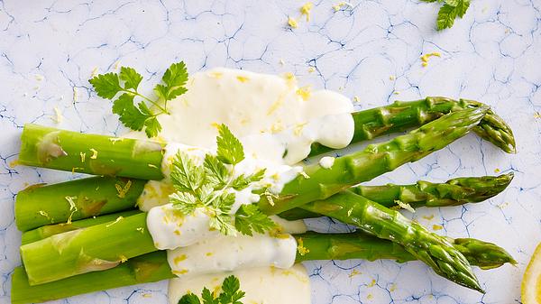 Schnelle Spargelsoße: Einige Stangen gegarter grüner Spargel liegen auf einem blau-weiß marmorierten Untergrund. Die Spargelstangen sind mittig mit einer hellgelben Soße bedeckt. Darauf befinden sich einige Fähnchen Kerbel sowie Zitronenzeste. - Foto: House of Food / Bauer Food Experts KG